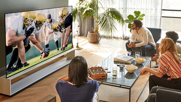 Um grupo de amigos sentados na sala de estar com um amplo ângulo de visão e a assistirem a um jogo de futebol na TV