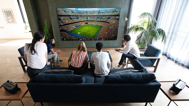Um grupo de amigos a assistirem a um jogo desportivo na TV na sala de estar com 2 colunas Bluetooth iguais na posição de colunas traseiras