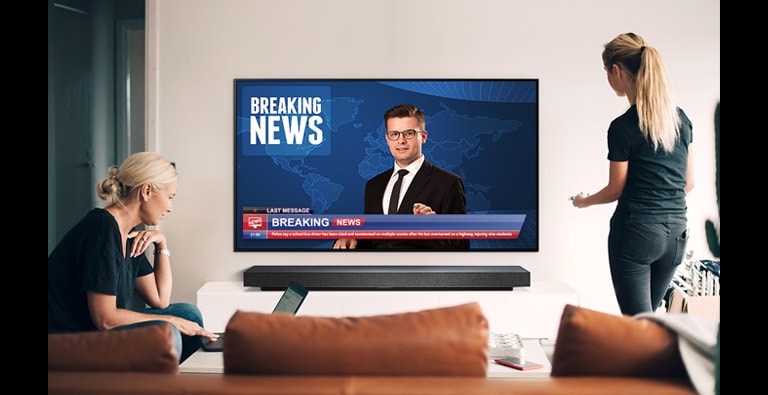 Uma mulher de pé a ver as notícias em alta qualidade na TV e outra sentada em frente ao portátil (mova o diapositivo)