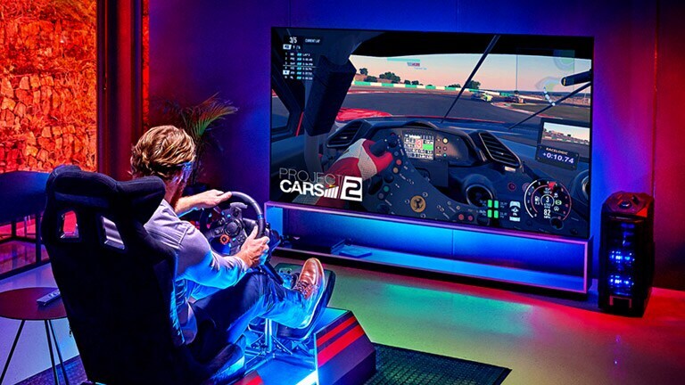 Homem numa sala escura, sentado numa cadeira de jogos de corrida à frente de uma TV grande a jogar num jogo de corrida