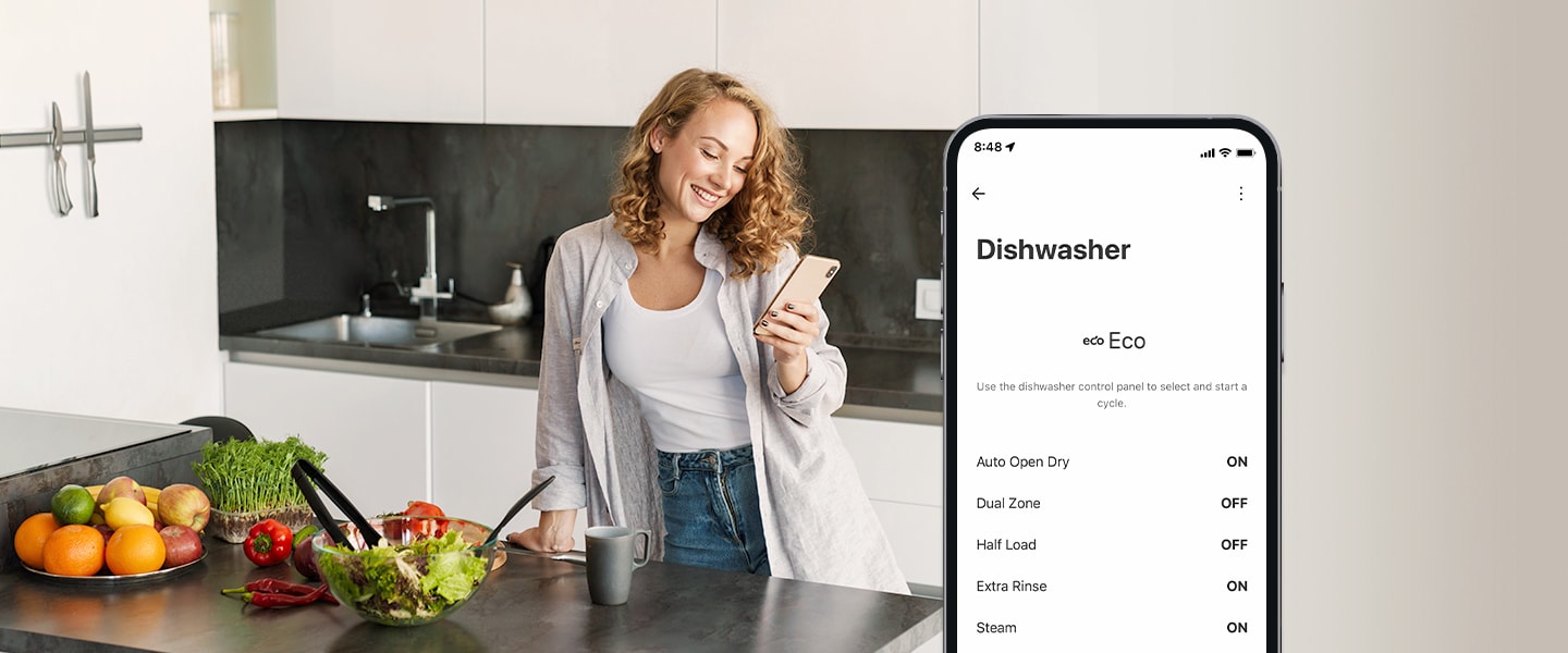 Uma mulher olha para o seu telemóvel na cozinha e o smartphone mostra as definições de personalização na aplicação ThinQ™.