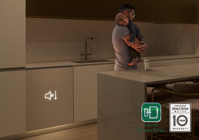 Homem segura num bebé a dormir numa cozinha pouco iluminada, máquina de lavar loiça de encastre trabalha no fundo.
