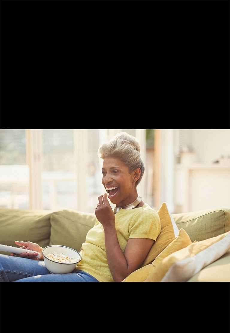 Uma mulher está a ver TV e a segurar um controlo remoto. Também está a comer pipocas.