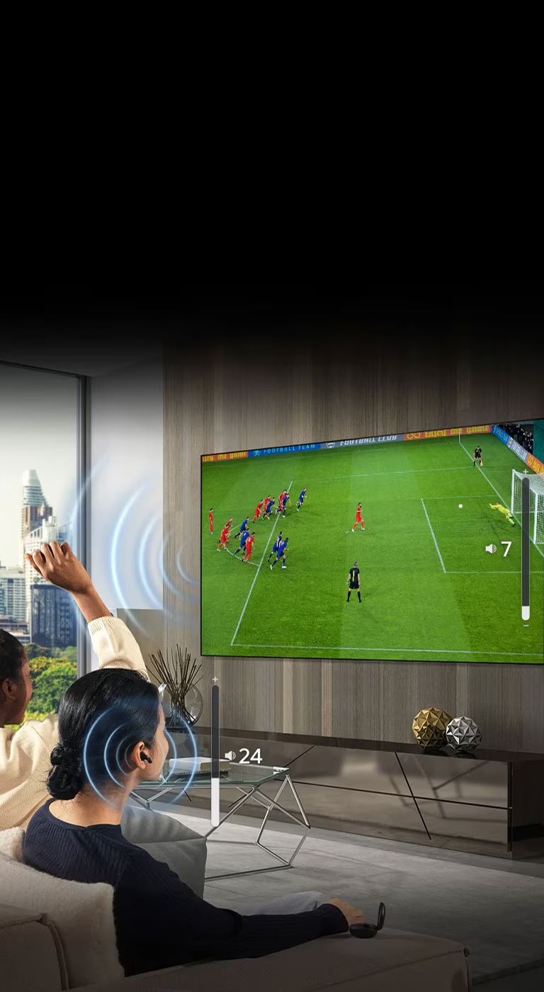  Um grupo de pessoas está sentado num sofá a ver um jogo de futebol na TV. A mulher na extremidade direita está a usar auriculares e usa-os com um volume diferente do volume da TV, indicando que está a usar ambos ao mesmo tempo.