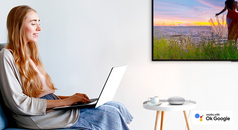 Uma mulher sentada num sofá com um portátil, numa divisão com uma TV numa parede, controla uma coluna AI para desativar a TV.