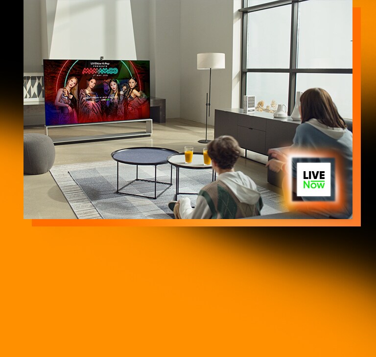 Duas pessoas sentadas na sala de estar a ver TV: uma imagem de teaser do mamamoo no ecrã da TV. Logótipo da Livenow colocado no canto inferior direito. 