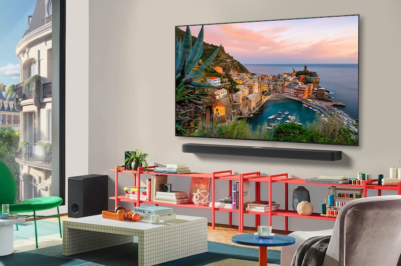 منظر واسع لغرفة معيشة أنيقة وملونة. تلفزيون LG OLED AI CS5 4K Smart TV مثبت على الحائط مع مكبر صوت LG Soundbar أسفله. يتناغم تصميم التلفزيون بشكل جيد مع المساحة. يوجد على شاشة التلفزيون عمل فني ملون. 