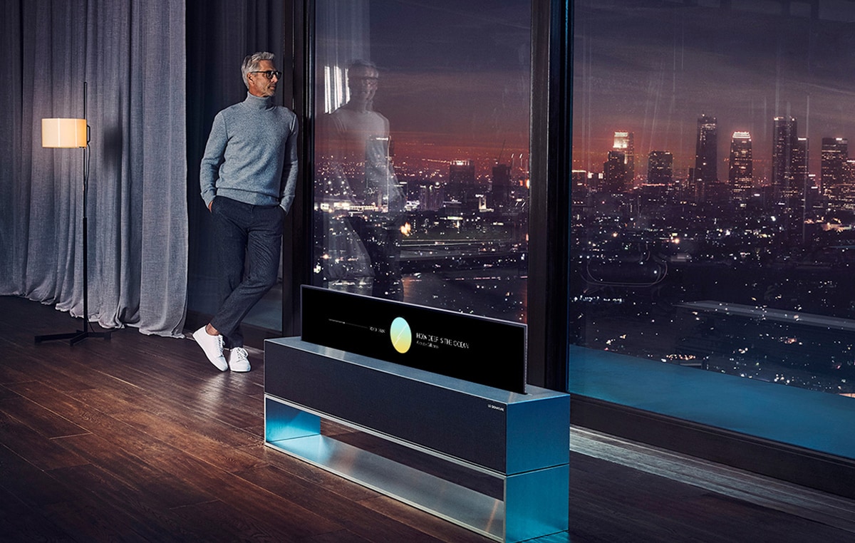 رجل يتكأ على نافذة غرفته الكبيرة ويُطالع منظر المدينة ليلاً وبجواره تلفزيون LG SIGNATURE Rollable OLED في وضع العرض الخطّي.