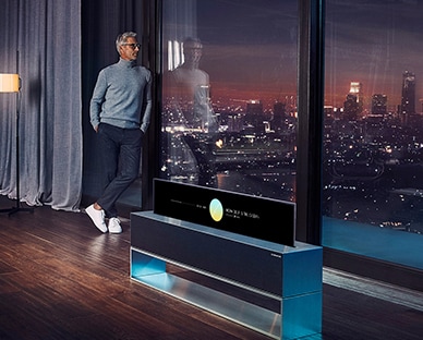  رجل يتكأ على نافذة غرفته الكبيرة ويُطالع منظر المدينة ليلاً وبجواره تلفزيون LG SIGNATURE Rollable OLED في وضع العرض الخطّي.