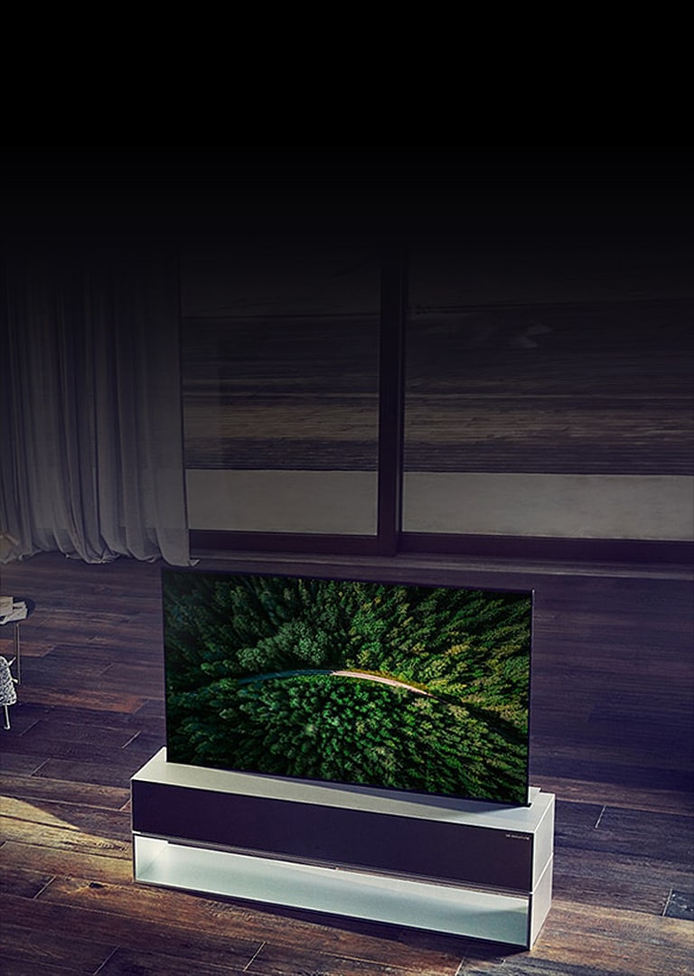 رجل يجلس في غرفة المعيشة ويشاهد تلفزيون LG SIGNATURE Rollable OLED موضوع على أرضية خشبية.