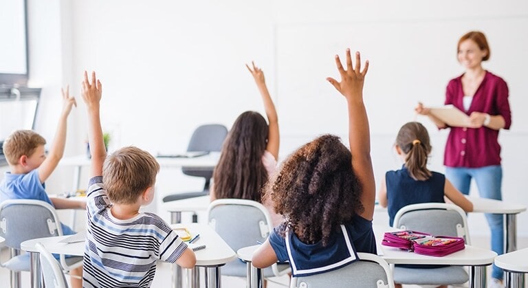 Image of a classroom with a teacher and studends raising their hands.