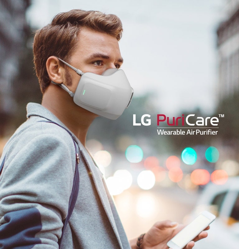 A man stands downtown in a city with the LG Puricare Mask on, holding a phone, and looking around with a blurred city in the background.