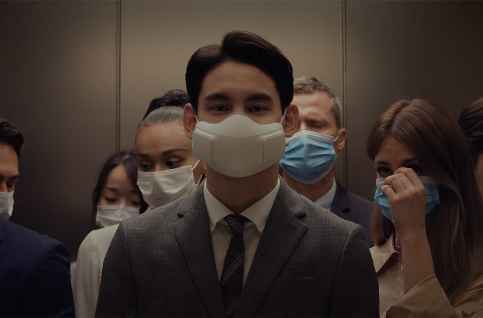 A video opens with a crowded elevator. The man in front is wearing the PuriCare Wearable Air Purifier while everyone else is wearing normal cloth masks except one person. The person not wearing a mask coughs and everyone else in the elevator moves uneasily while the man with the PuriCare mask stands confidently and a script comes into the center reading "Air doesn't always have to be shared". The camera moves in to zoom in on the mask and then goes into the filter and shows a magnified focus on the inner workings of the mask. The bottom reads "The PuriCare Filter blocks out dust, viruses, bacteria and allergens". The focus goes through to the person's mouth and shows a mouth breathing comfortably and then back out through the filter to see the crowded elevator again. Next a white background and then the words "Technology that lets you breath easy". Finally the words disappear and the LG PuriCare Wearable Air Purifier logo ends the video.