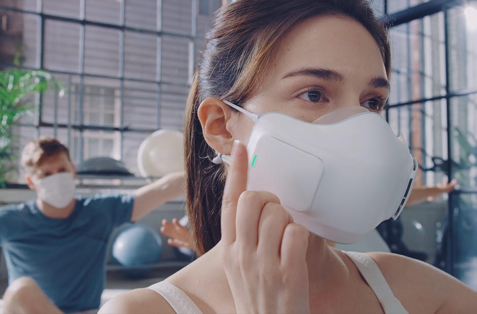 A video opens with a yoga class. One of the people in the class wears the PuriCare Wearable Air Purifier while the rest of the students wear regular cloth masks. The students stretch up and the camera focuses on a man wearing a normal mask who moves his mask below his nose and mouth as he struggles to breathe and work out. The background blurs and the words "Breathing shouldn't be an exercise in frustration" appear. Next focus moves to the woman wearing the PuriCare mask. As she begins to breathe harder, she taps the side of the mask twice to increase air filtering. The mask becomes invisible with only the fans showing to show how she can easily breathe through the mask even while working out. The words "DUAL Fans activate in sync with your breathing to help take in air" appear at the bottom. As the woman inhales, the fans glow blue and as she exhales the fans go dark easily keeping up with her. The students lie down to stretch and the zoom pulls out showing her PuriCare mask once again and all of the other students wearing cloth masks. Next a white background and then the words "Technology that lets you breathe easy". Finally the words disappear and the LG PuriCare Wearable Air Purifier logo ends the video.