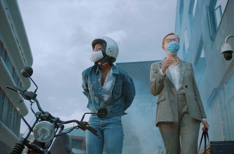 A video opens with a woman disembarks from her motorcycle in a busy city. She's wearing the PuriCare Wearable Air Purifier and pedestrians passing are wearing normal cloth masks. A man passes by with the cloth  mask beneath his chin and a woman with a cloth mask walks up and stops. Her glasses have fogged up and she takes them off and stretches her neck and takes off her cloth mask to relax her ears. The background blurs and the words "All day discomfort doesn't have to be your new normal" appear. The focus goes back to the woman in the PuriCare mask who takes off her helmet. The camera focuses on the comfort aspects of the mask. The mask goes invisible and the Face Guard can be seen around the woman's mouth. The words "Flexible Face Guard hugs the contours of your face while minimizing air leakage" appears on the bottom. The camera moves down to show the chin vent. The words "Easy exhalation vent and soft ear straps provide all day comfort" appear on the bottom. The focus moves to the ear straps resting comfortably around the woman's ears. The camera moves out to show the woman wearing glasses that haven't fogged and the mask is not invisible anymore. Next a white background and then the words "Technology that lets you breath easy". Finally the words disappear and the LG PuriCare Wearable Air Purifier logo ends the video.