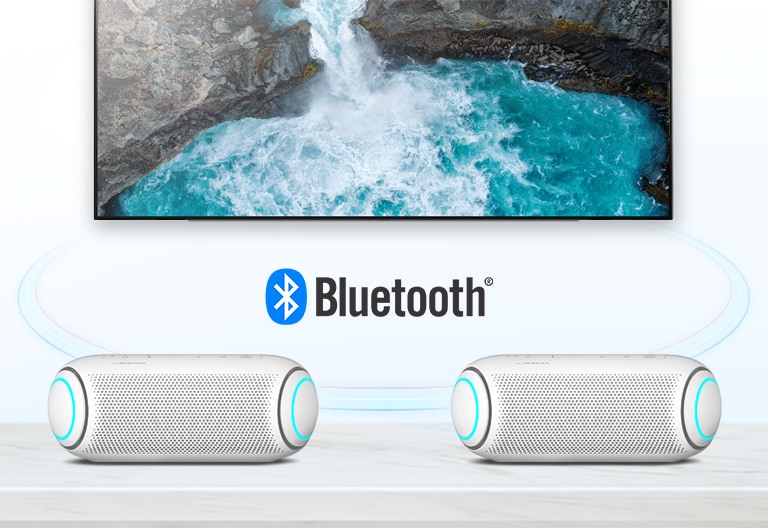 On a table, two LG XBOOM Go with sky blue lighting are in front of a TV showing a waterfall.