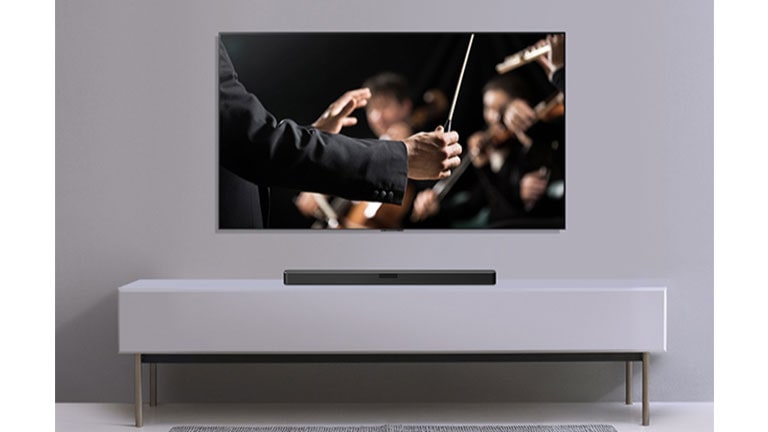 A TV is shown on a gray wall and LG Soundbar below it on a gray shelf. TV shows a conductor conducting an orchestra.