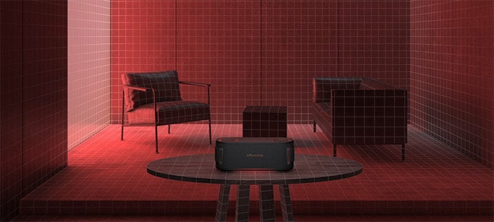 In a room filled with a red lighting a black sofa, a black ottoman  and a black armchair are placed behiind. In the fornt, xboom Bounce is placed on the top of black round table.	