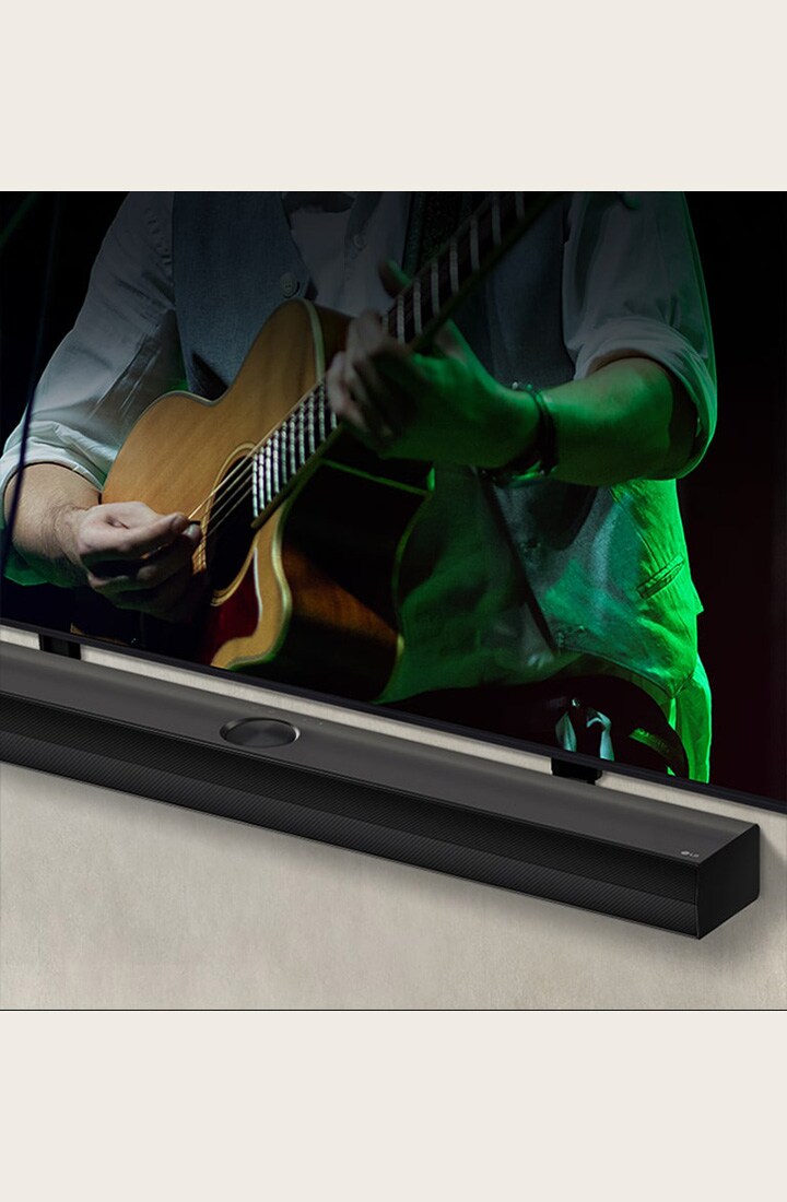 LG Soundbar against a black backdrop reveals its design starting from the left corner, then pans out to show the whole soundbar. An LG QNED TV appears with Synergy Bracket. The Soundbar sits on top of the Synergy Bracket, presses against the wall with the lower screen of the TV being visible, displaying a man playing the guitar.