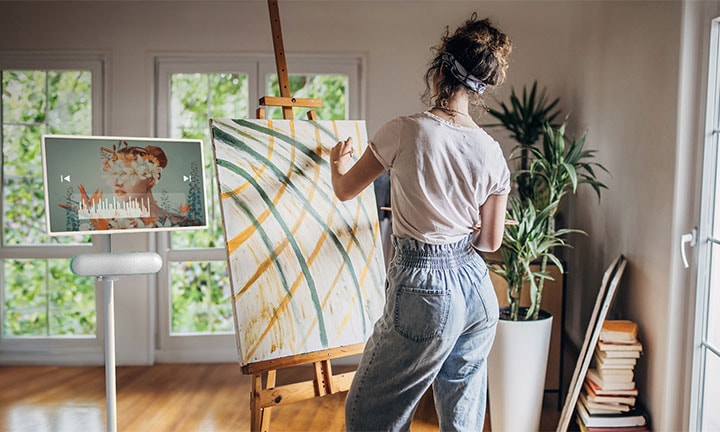 The LG StanbyME is placed in the livingroom with the XT7S speaker attached. A woman draws while listening to music through the screen and speaker combo.