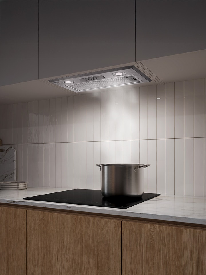 Built-in hood in a dim kitchen gently pulling steam upward from a pot on the cooktop.