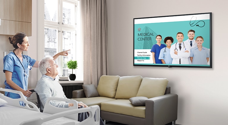 A TV is installed on the hospital room’s wall, and a nurse points to it while explaining to the patient.
