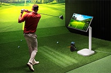 A man is playing golf at an indoor screen golf facility, with a LG Smart Monitor Swing next to him displaying data.