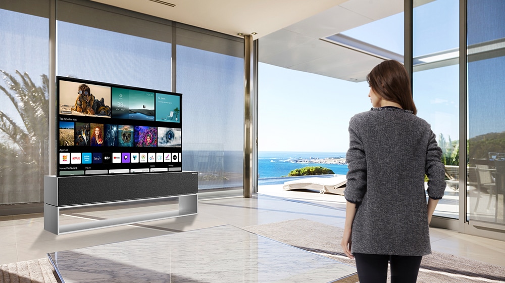 A woman standing in a living room is watching a TV displaying New Home screen.