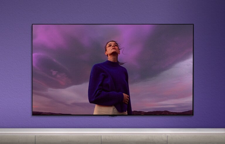 QNED TV is placed on a purple wall and the screen shows a woman wearing a purple shirt. 