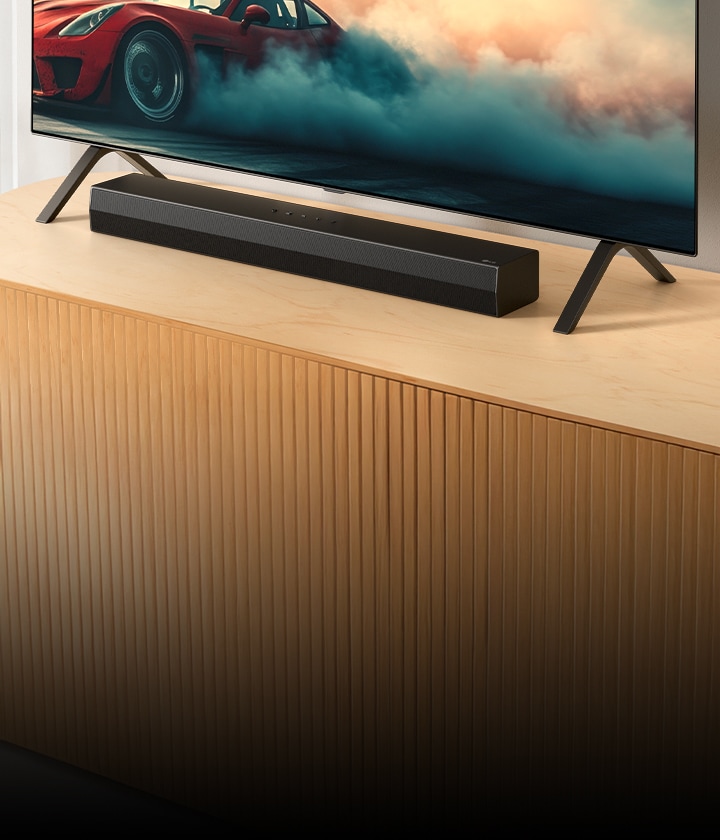 LG Soundbar S20A is placed on a wooden TV console, below a TV and between its TV stands. On the TV a red car with smoke is shown.