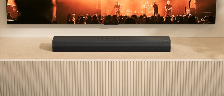 S20A soundbar is placed on a wooden TV console. Above it a TV is partially shown with a band's concert scene on it.