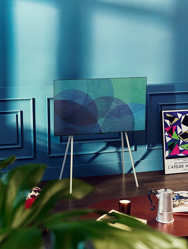 A floor stand LG OLED television is in a modern gray room in front of a window that looks onto a snowy forest. A floor stand LG OLED television is in a colorful room with pastel and wooden furnishings.