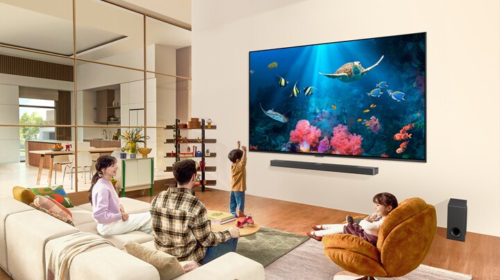 En familj i ett vardagsrum med en ultrastor LG-TV monterad på väggen, en havsscen med koraller och en sköldpadda på skärmen.