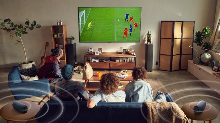 Fem personer som sitter i en soffa tittar på en storbilds-TV som visar en fotbollsmatch, med Bluetooth-högtalare som ger surroundljud.