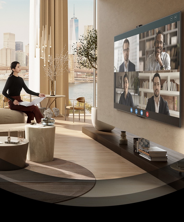En kvinna sitter i en beige soffa i ett rymligt vardagsrum och håller i en bärbar dator och har ett videosamtal på en stor TV.