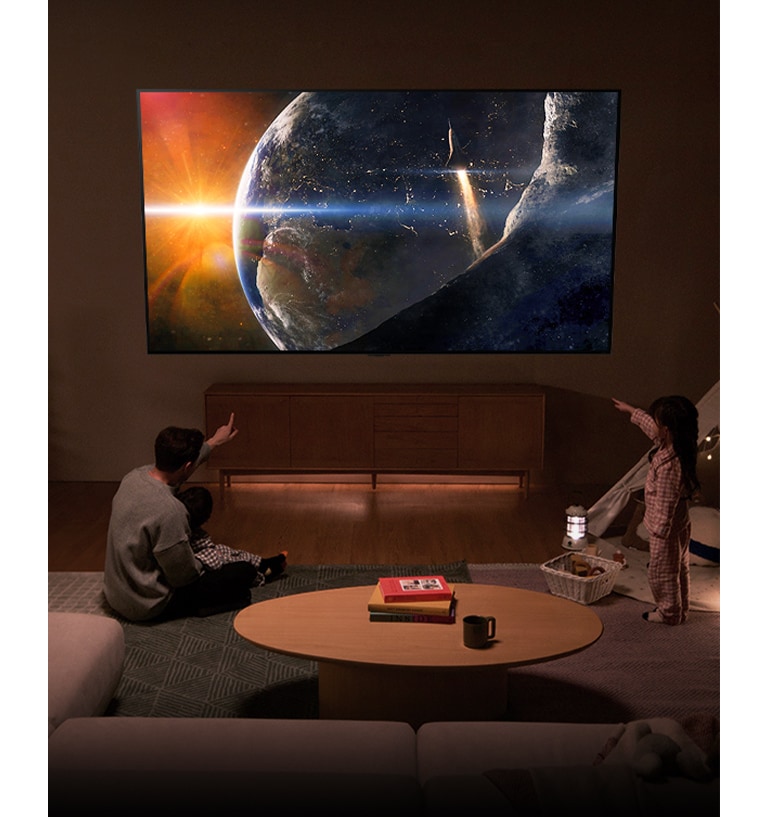 En familj sitter på golvet i ett lågt upplyst vardagsrum vid ett litet bord och tittar upp på en LG-TV monterad på väggen som visar jorden från rymden.