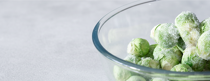 A glass bowl filled with frozen Brussels sprouts lightly covered in frost, placed on a light gray surface.