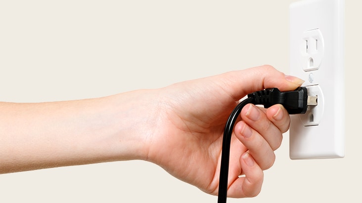 A hand plugging a black power cord into a white electrical outlet on the wall.