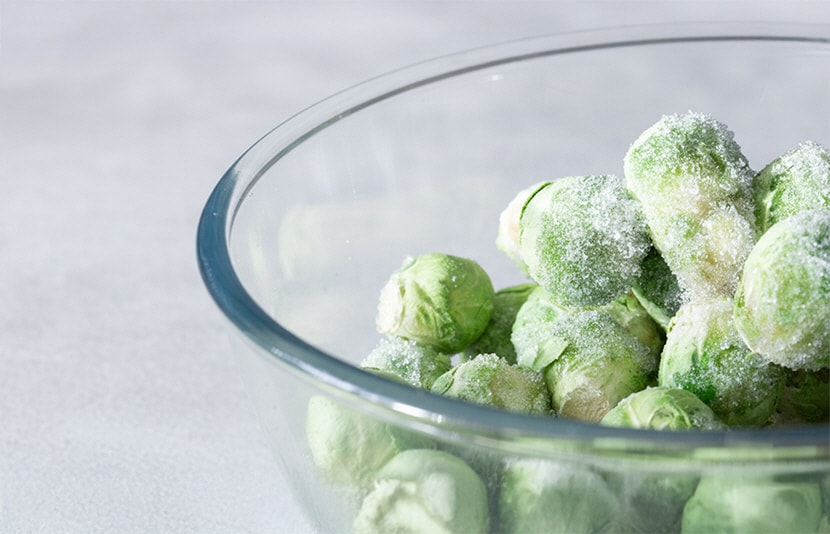 A glass bowl filled with frozen Brussels sprouts lightly covered in frost, placed on a light gray surface.