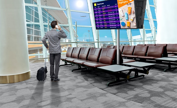 A traveler stands in an airport lounge, checking his phone while looking at a digital departure board displaying flight schedules. The scene highlights the modern, efficient use of signage for real-time updates.
