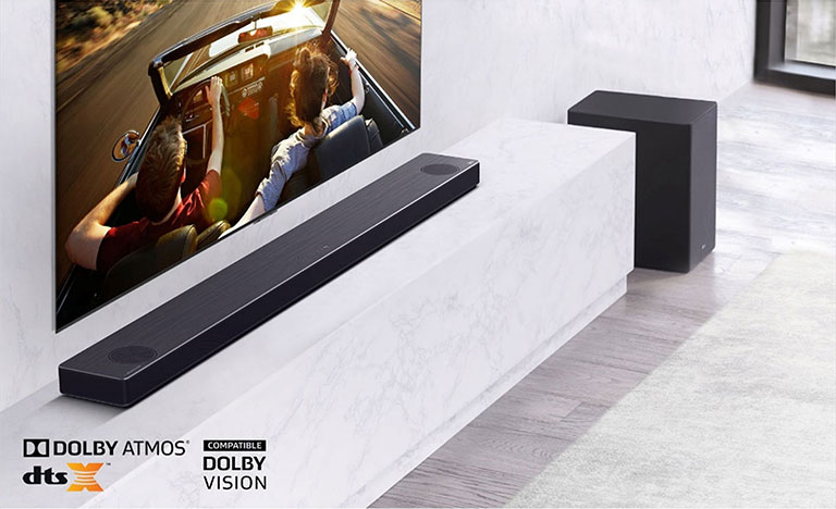 TV is on the wall, LG Soundbar is below on a white marble shelf with a sub-woofer to the right. TV shows a couple in a car.