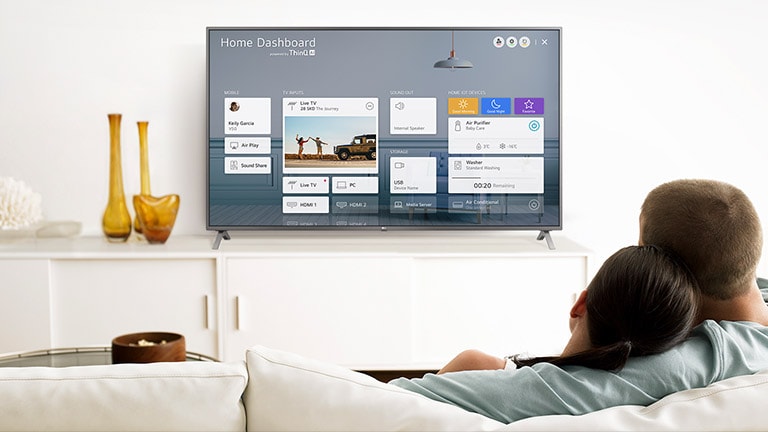 A men and women sitting on a sofa in the living room with the Home Dashboard on the TV screen