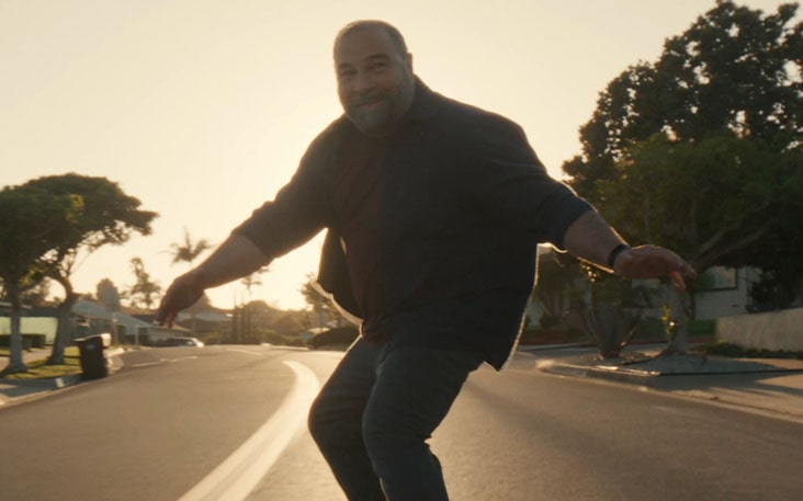 A middle-aged man riding a longboard with bright sunlight shining behind.