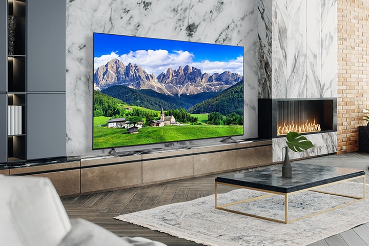 A large TV is placed on a living room wall decorated with a fireplace. The scenery of a mountain and a village is bright and vivid on the TV screen.	