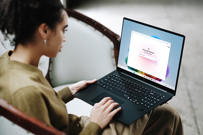 This image shows a woman sitting on a chair using a laptop. The screen displays an AI-powered search interface with a search bar, suggesting intelligent assistance or voice search functionality.