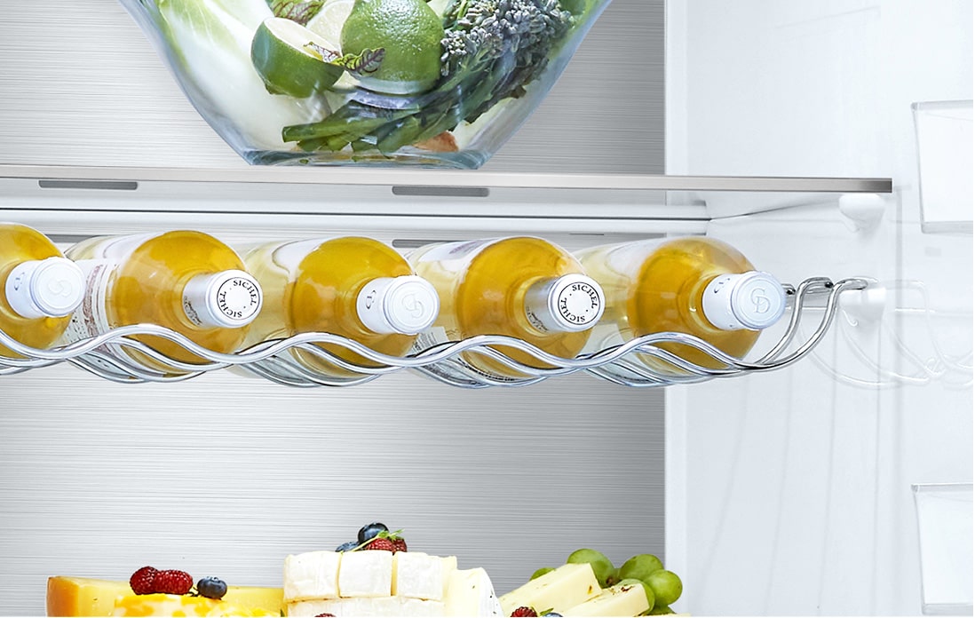 Inside a fridge, wine bottles lie on a metal rack beneath a shelf with a salad bowl, and cheese with fruits below.