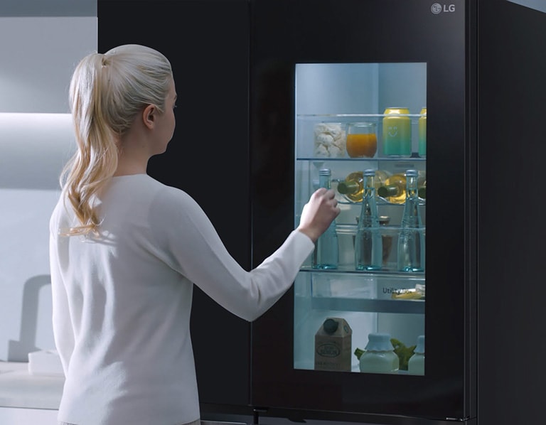 A video shows a woman approach her InstaView refrigerator and knock twice. The interior lights up and she can see the contents of her fridge without opening the door. The view zooms in to focus on the drinks in the door and then zooms out to see the woman from behind as she opens the door and grabs a drink.