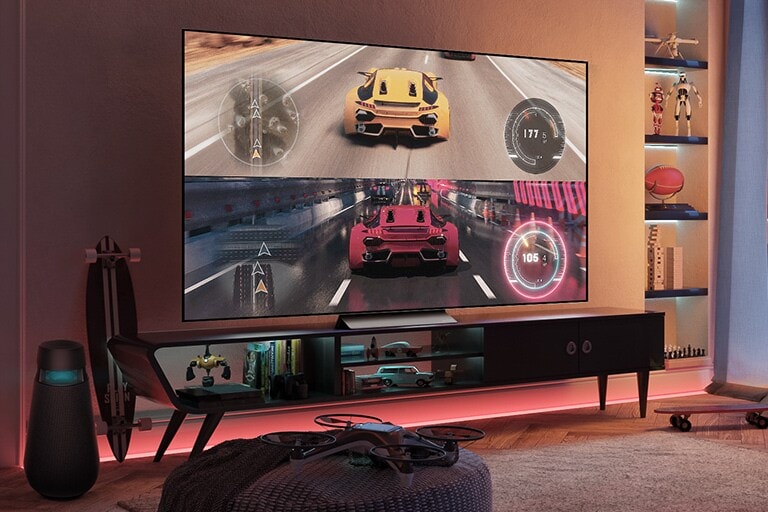 In a game room with red lighting, a racing game image of yellow and red cars driving is displayed on a large screen TV. On the left side, there is a dedicated game speaker, and there are gaming and device-related accessories around.