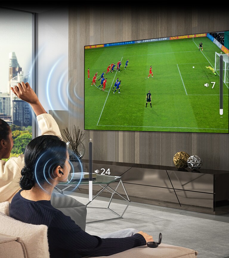 A group of people are sitting on a sofa watching a soccer game on TV. The woman on the far right is wearing earbuds and using them with a different volume than the TV, indicating that she is using both at the same time.