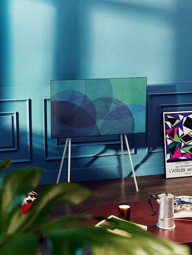 A floor stand LG OLED television is in a modern gray room in front of a window that looks onto a snowy forest. A floor stand LG OLED television is in a colorful room with pastel and wooden furnishings.